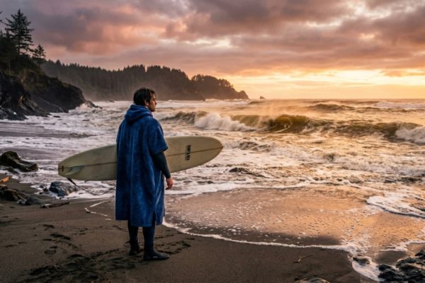 Surfer wearing a warm hooded surf poncho on a chilly California beach at sunset.